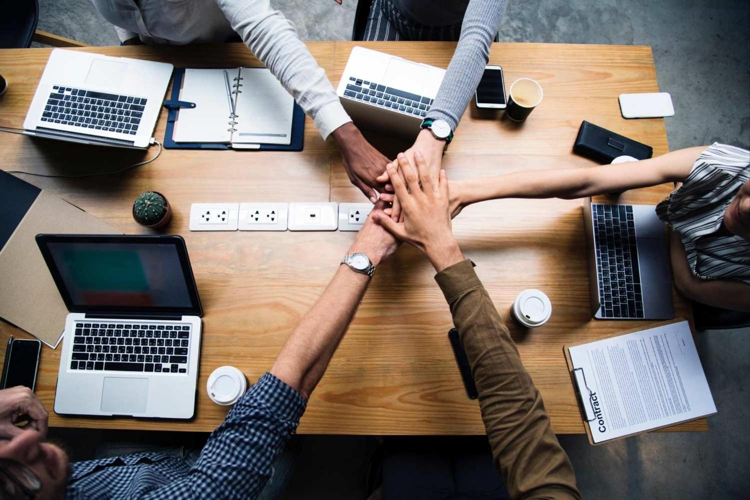 A team gathered around a table with laptops and notes, hands joined in the middle